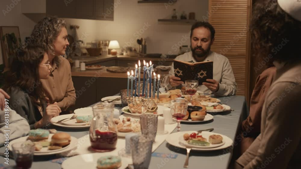 Medium full shot of middle-aged Jewish man in yarmulke sitting at Hanukkah feast with family, reading text from holy Torah with star of David on cover, and candles burning on hanukkiah on table Medium full shot of middle-aged Jewish man in yarmulke sitting at Hanukkah feast with family, reading text from holy Torah with star of David on cover, and candles burning on hanukkiah on table