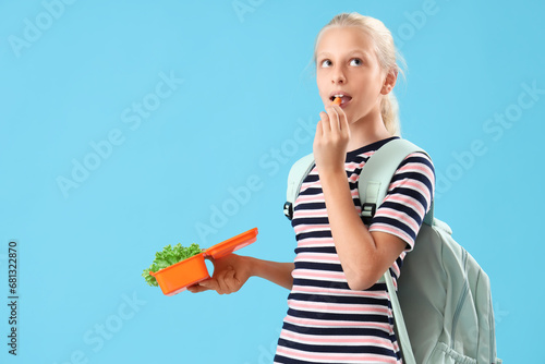 Thoughtful girl with backpack and lunchbox eating vegetables on blue background