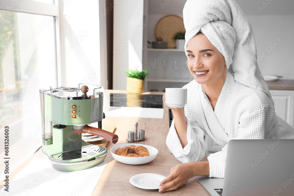 Beautiful young woman in bathrobe drinking coffee and using laptop at kitchen Beautiful young woman in bathrobe drinking coffee and using laptop at kitchen