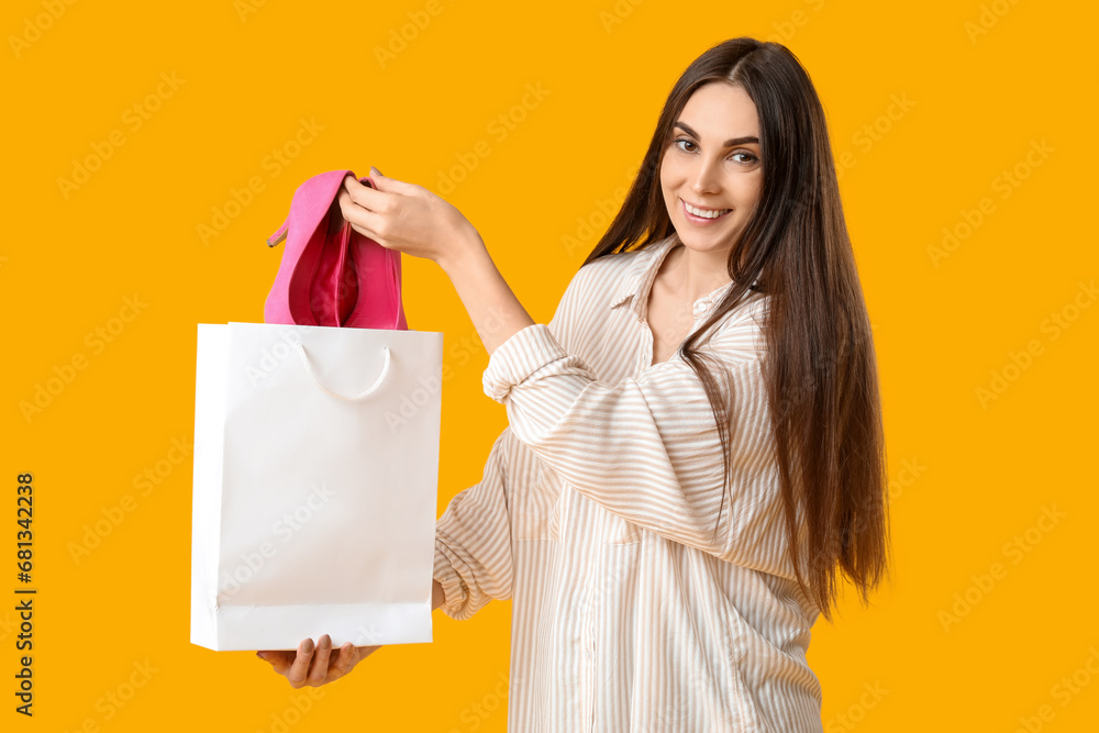 Beautiful young woman holding shopping bag with stylish pink high heels on yellow background Beautiful young woman holding shopping bag with stylish pink high heels on yellow background