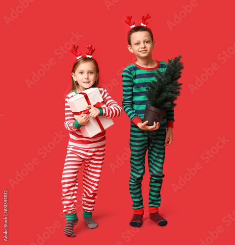 Cute little children in pajamas with Christmas gift and tree on red background