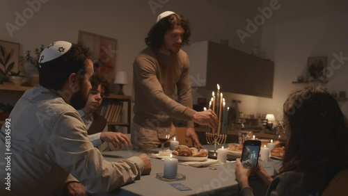 Medium shot of widowed middle-aged Jewish man in kippah celebrating Hanukkah at home with his children, eldest son lighting up candles on menorah and young daughter filming on smartphone photo