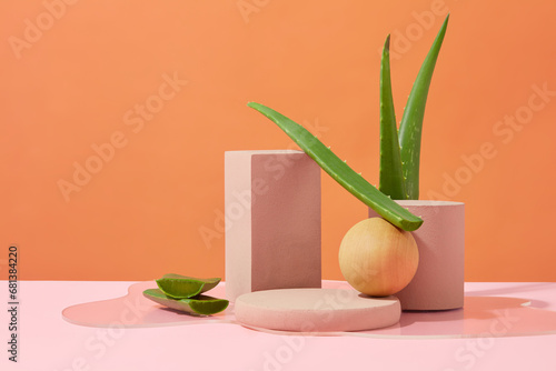 Platforms of different shapes are arranged next to each other on a pink table. Orange background with aloe vera leaves. Ideal space for advertising cosmetics with aloe vera extract.