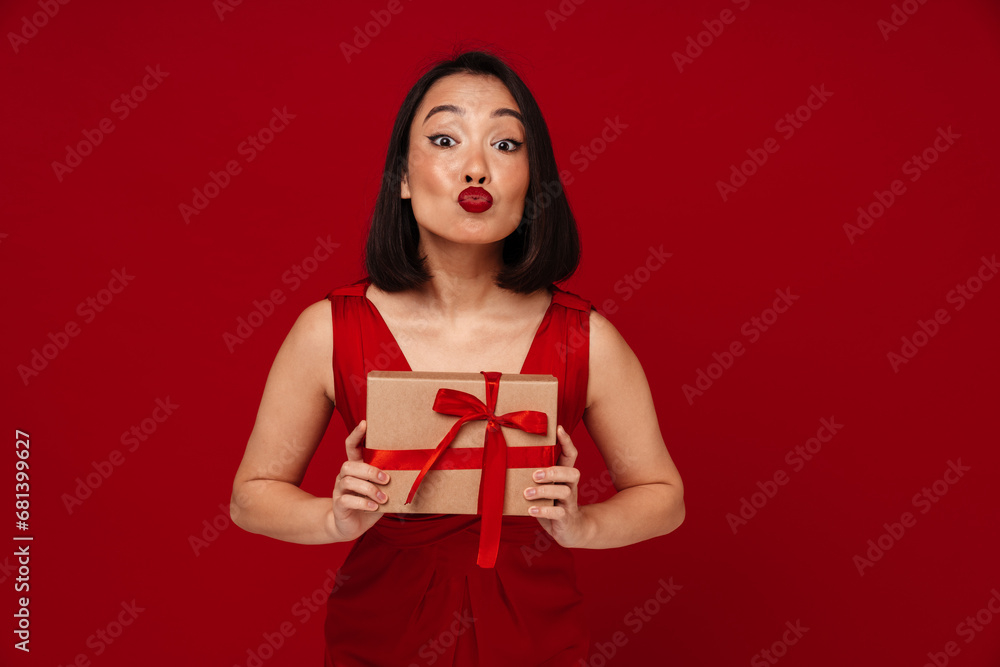 Fototapeta premium Young woman holding gift box and making kiss lips isolated over red background