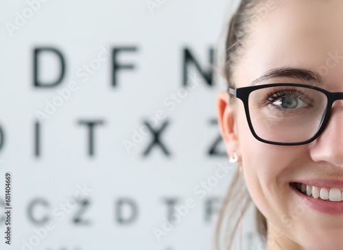 Focus on beautiful woman wearing glasses in black stylish frame. Cheerful ophthalmologist looking at camera with happiness. Letters table for eyesight check on background. Vision test concept