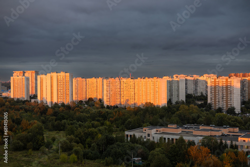 Tall multi-storey buildings in the rays of the setting sun. A beautiful residential area of the city at sunset against a cloudy sky. Multi-storey residential buildings in an urban area at sunset. © Александр Лебедько