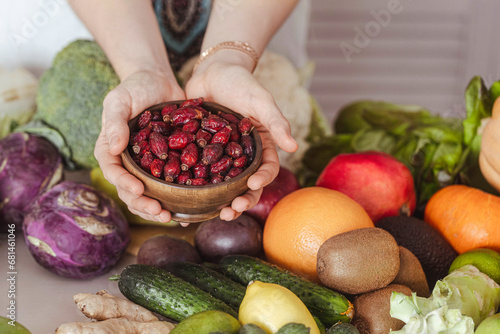 Female hands with dried rosehip in wooden bowl above vegetables and fruits on a table in the kitchen, close-up, selective focus. photo