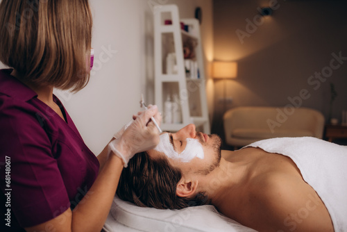 Fotografie Man getting facial nourishing mask by beautician at spa salon, closeup