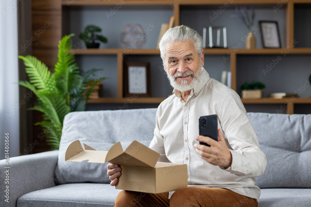 Portrait of a smiling gray-faced senior man who is happy to shop online, holds a delivered package, a mobile phone and smiles at the camera Portrait of a smiling gray-faced senior man who is happy to shop online, holds a delivered package, a mobile phone and smiles at the camera