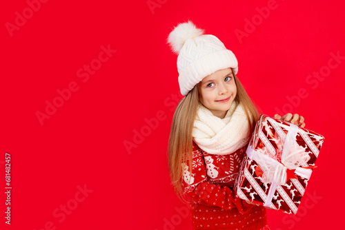 a child girl in a winter hat and sweater with gifts on a red monochrome isolated background rejoices and smiles  the concept of new year and Christmas  space for text