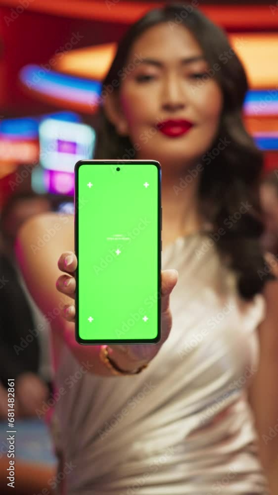 Vertical Screen: Advertising Template with an Asian Female Showing a Smartphone with a Green Screen Mock Up Display. Professional Beautiful Model in a Casino with People Gambling in the Background Vertical Screen: Advertising Template with an Asian Female Showing a Smartphone with a Green Screen Mock Up Display. Professional Beautiful Model in a Casino with People Gambling in the Background