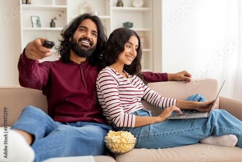 Relaxed millennial couple chilling at home, watching tv, using laptop