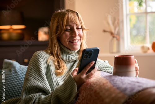 Woman At Home With Warming Hot Drink Of Tea Or Coffee In Cup Or Mug With Mobile Phone Streaming