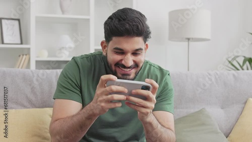 Bearded indian man winning in video games on modern smartphone while sitting on comfy couch at home. Young millennial guy in green t-shirt making yes gesture and sincerely rejoicing. photo