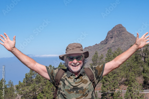 Happy joyful senior man in Tenerife carrying backpack enjoying trekking day on the top of mountain, elderly male outstretched arms looking at camera