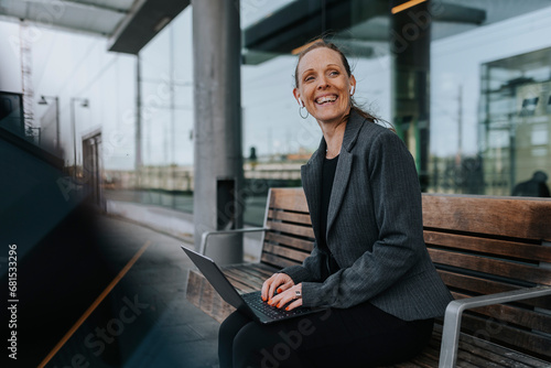 Cheerful businesswoman sitting with laptop on bench while waiting at railroad station