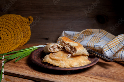 Clay plate of fried meat pies with cutlery and green onion on wooden table.