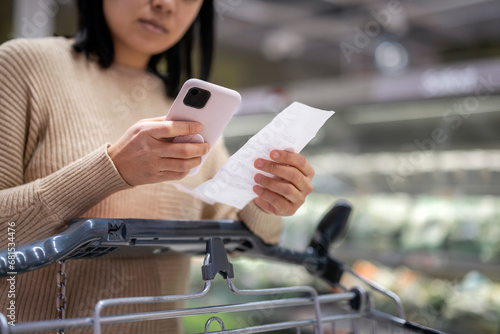 Young woman examining list while using smart phone at supermarket photo