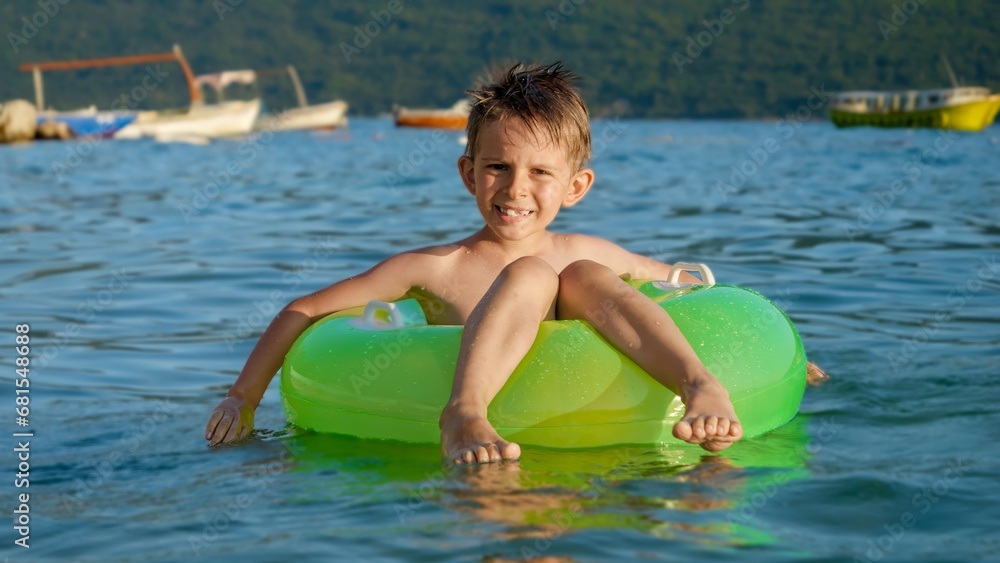 Happy cheerful boy splashing water while sitting in inflatable ring and swimming on calm sea waves at sunset. Family holidays, vacations, summertime.