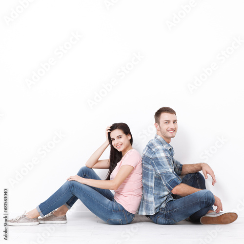 A frontal view captures the joyous moment of a young couple seated on the floor, backs turned to each other, both looking at the camera with big smiles isolated on white. 