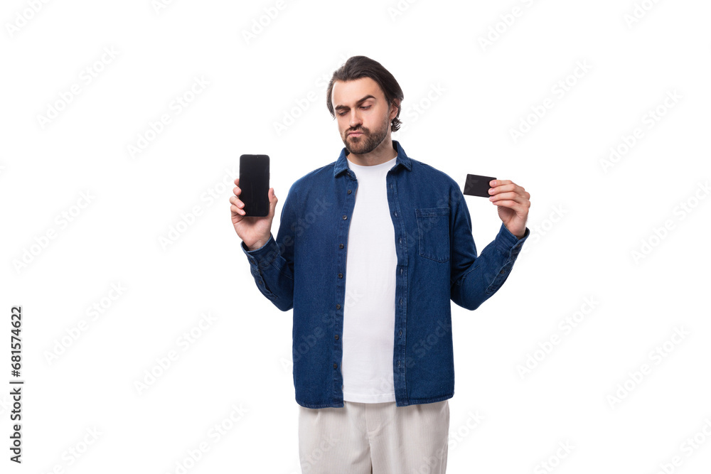 young caucasian man with black hair demonstates a plastic card and a smartphone. e-business concept young caucasian man with black hair demonstates a plastic card and a smartphone. e-business concept