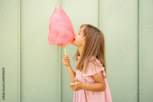 Portrait of a little girl with pink cotton candy on green wall background outdoors. Happy kids visiting amusement park and having fun photo