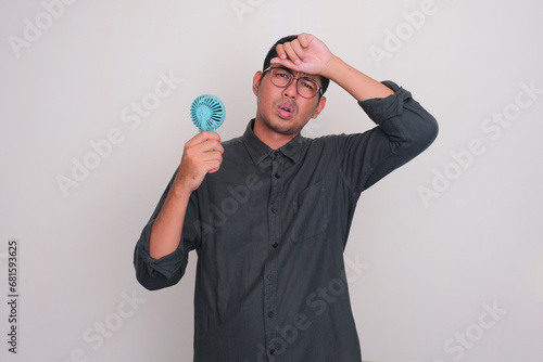 Adult Asian man using electric hand fan and showing swelter gesture photo