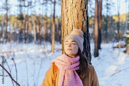 Attractive brunette female nature lover with back pressed against a tree at pine forest during the winter time.