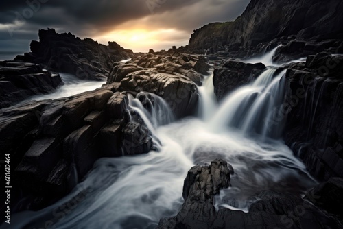  a long exposure photo of a waterfall in the middle of a rocky area with a dark sky in the background and the sun peeking through the clouds in the distance.