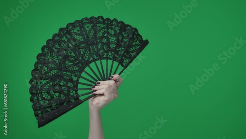 In the frame on a green background, a chrome key female hand with a manicure holds a black, openwork fan. She opened it, used to inflate the air. Here can be your advertising. Medium frame photo