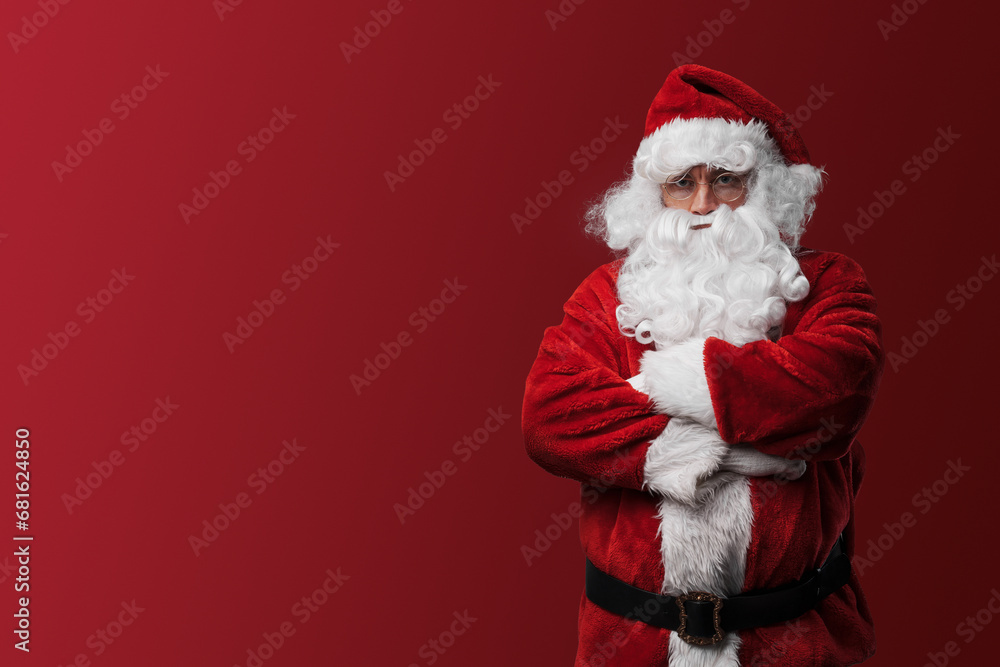 Serious Santa Claus in red costume posing with crossed arms against a red background Serious Santa Claus in red costume posing with crossed arms against a red background
