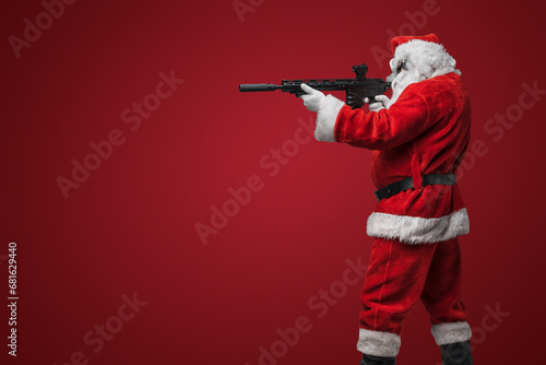 A man dressed as Santa Claus, wearing sleek black sunglasses, poses with toy machine guns against a bold red backdrop photo