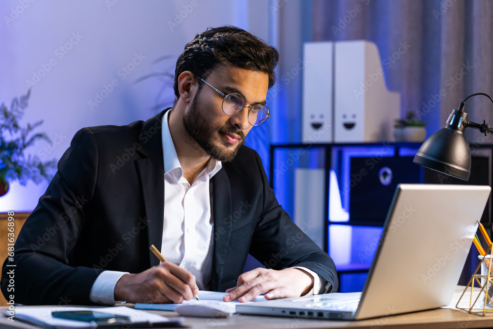 Focused serious Indian businessman freelancer working at modern home office workplace desk writing notes ideas in notebook. Male Hindu in formal suit preparing planning presentation startup project