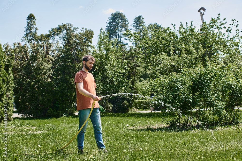 bearded gardener in casual attire holding gloves and watering green lawn while working outdoors bearded gardener in casual attire holding gloves and watering green lawn while working outdoors