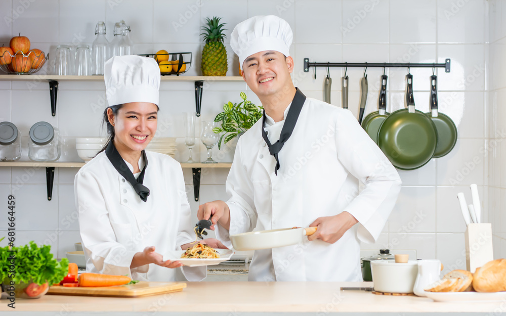 Two Asian professional couple chef wearing white uniform and hat, helping for preparing ingredients for healthy meal, cooking in kitchen together, smiling with happiness and confidence. Food Concept.
