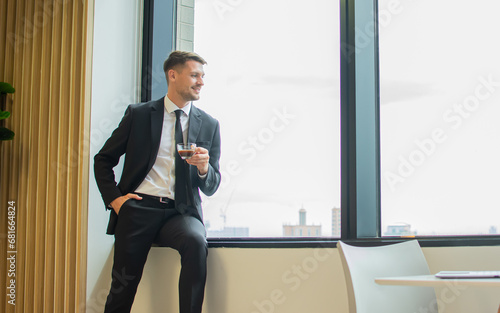Handsome smart business man wearing formal black suit and necktie, holding cup of coffee, smiling with happiness, standing and looking outside window in morning, taking break in indoor office.