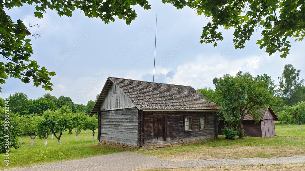 An old wooden house with shingle roof and lightning rod is located among the trees near an orchard with whitewashed trunks. There is a tiled path to the house through a grassy lawn. Cloudy weather An old wooden house with shingle roof and lightning rod is located among the trees near an orchard with whitewashed trunks. There is a tiled path to the house through a grassy lawn. Cloudy weather