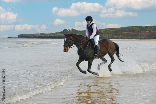 Young female rider and her bay horse pony gallop through waves at seas edge in Wales UK, splashing water and having a wonderful exciting time.