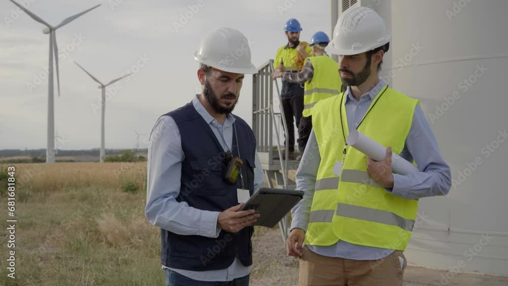 Team engineers men discuss maintenance of windmill in safety clothing using tablet at wind power plant in countryside. Mills and workers in background. Renewable energy clean sustainable future. Team engineers men discuss maintenance of windmill in safety clothing using tablet at wind power plant in countryside. Mills and workers in background. Renewable energy clean sustainable future.