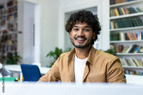 Close-up portrait of a young Muslim male student studying at a laptop in a university library, looking and smiling at the camera.