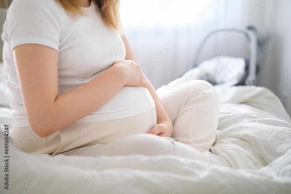 A soon to be mother, dressed in light attire, sits on the bed and lovingly cradles her pregnant belly. A soon to be mother, dressed in light attire, sits on the bed and lovingly cradles her pregnant belly.