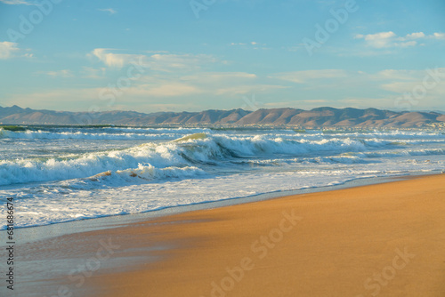 Fototapeta Naklejka Na Ścianę i Meble -  Light airy seascape. Wide sandy beach, sea waves, silhouette of mountains, and cloudy sky