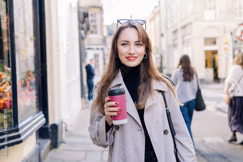 Young smiling woman in trench coat enjoying hot coffee drink in reusable cup while walking on the city streets decorated for the autumn holidays. Happy fall cozy mood, seasonal fashion style trend.
