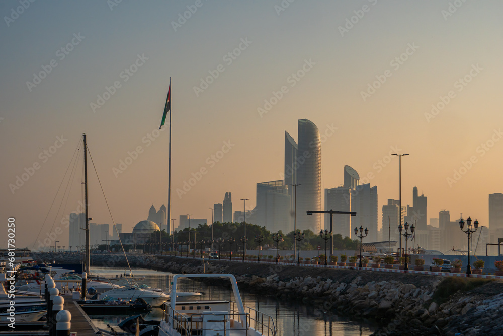 Naklejka premium Abu Dhabi, UAE, skyline from across the Corniche with a harbour, at sunrise. Tall UAE flag in the foreground.