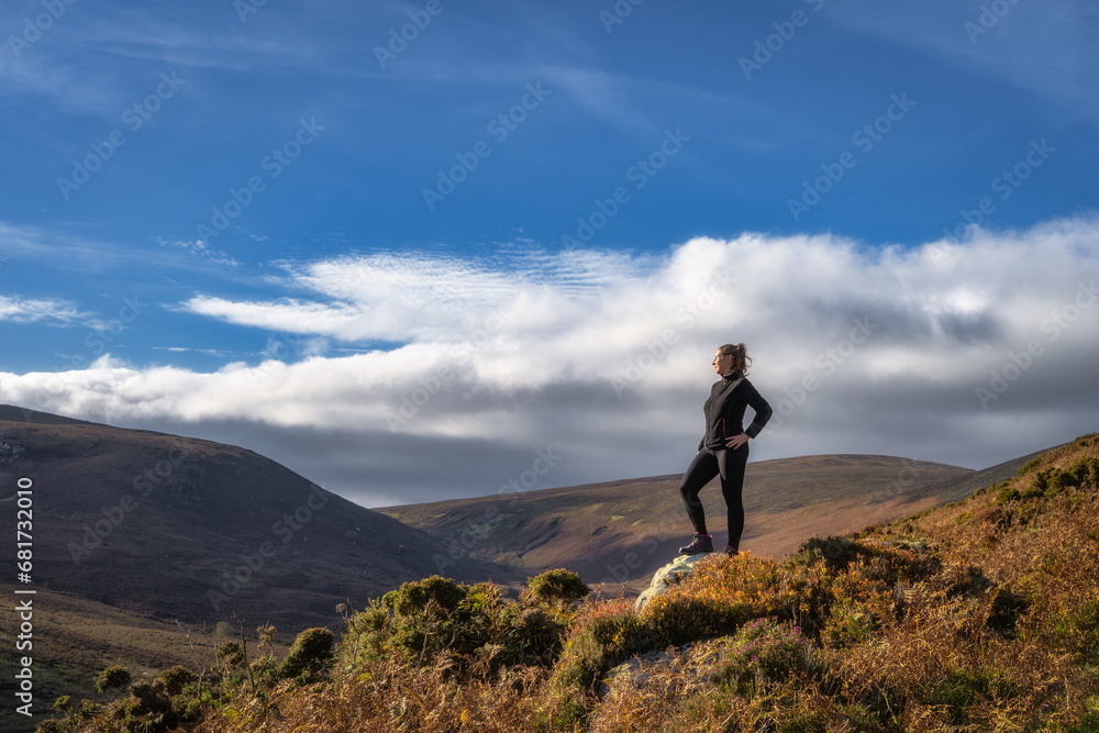 Adult woman standing on the rock, illuminated by sunlight, and looking at distant mountains. Hiking in Wicklow Mountains, Ireland