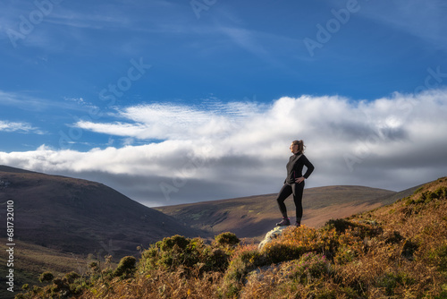 Fototapeta Naklejka Na Ścianę i Meble -  Adult woman standing on the rock, illuminated by sunlight, and looking at distant mountains. Hiking in Wicklow Mountains, Ireland