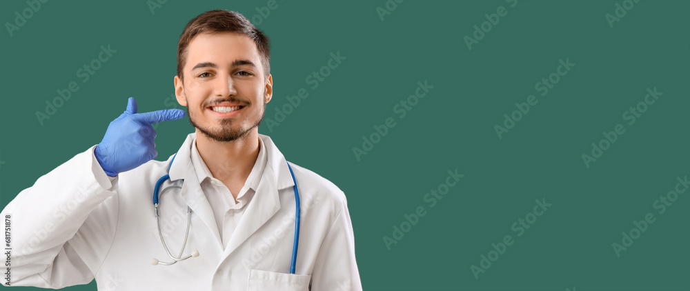 Young male dentist pointing at his teeth on green background with space for text