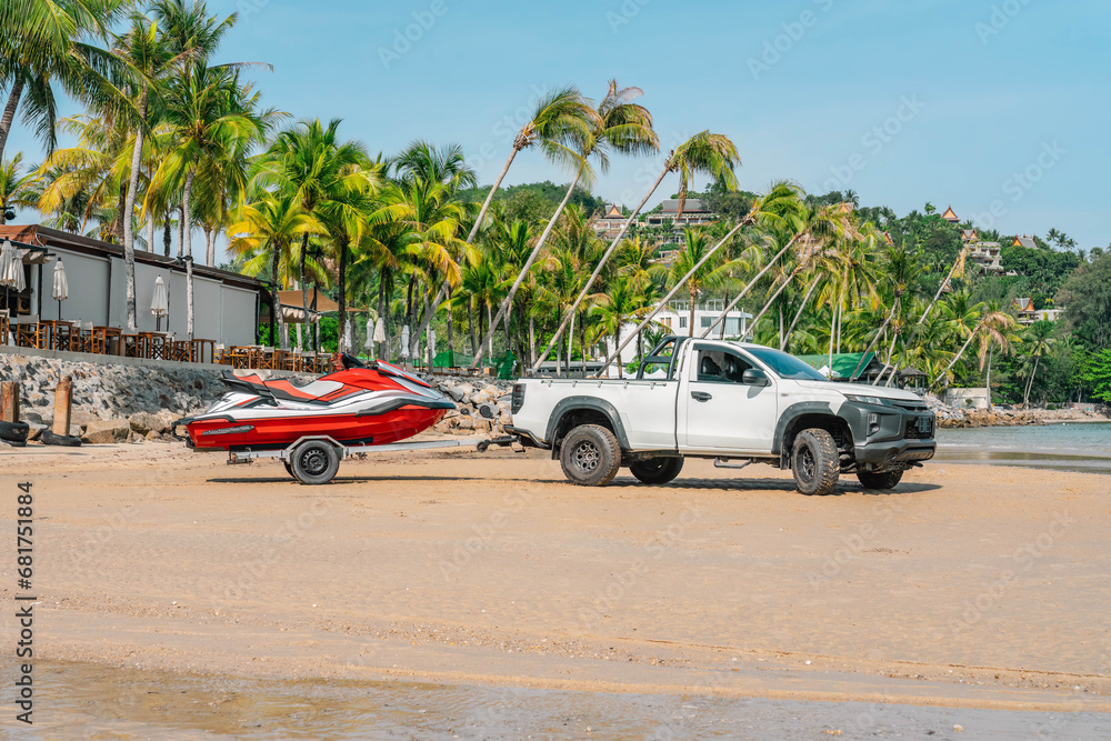 White pickup with jetski on trailer stands on sand beach, close to restaurant terrace and palm trees, morning time, tropical resort, Thailand - obrazy, fototapety, plakaty 