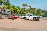 White pickup with jetski on trailer stands on sand beach, close to restaurant terrace and palm trees, morning time, tropical resort, Thailand
