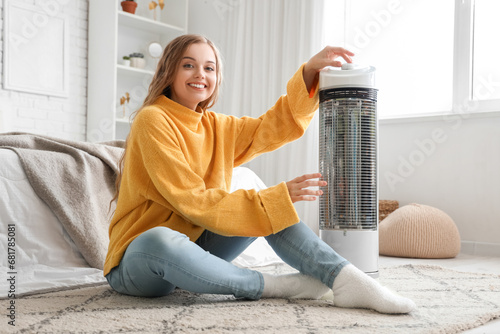 Young woman with electric heater sitting in bedroom